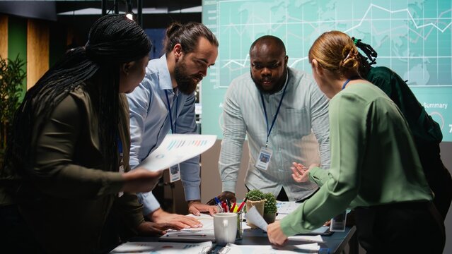 Multiethnic team exchanging files and review forecasting diagrams at night, meeting in a quiet office space. After hours collaboration to prepare key project updates for shareholders. Camera A.