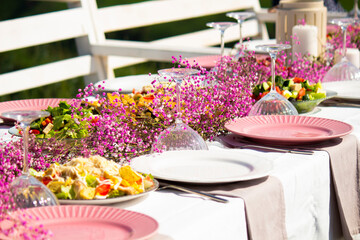 Outdoor festive table decorated with pink flowers and assorted food. Fresh vegetable salad with tomatoes and lettuce, elegant outdoor dining setup for celebration or wedding reception.