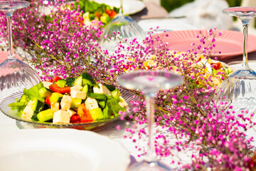 Close-up of festive table with pink flowers and assorted food. Fresh vegetable salad with tomatoes and lettuce, elegant outdoor dining setup for celebration or wedding reception.