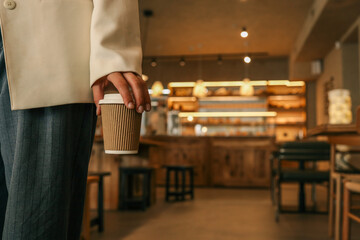 Close-up of hand holding takeaway coffee cup in stylish cafe interior, person wearing elegant...