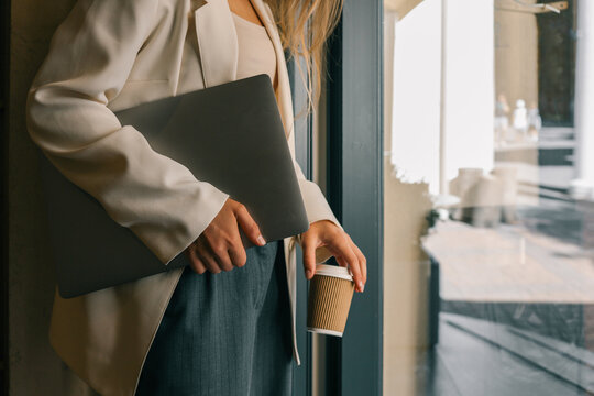 Businesswoman in cream blazer holding laptop and takeaway coffee cup near glass door, modern lifestyle and work concept
