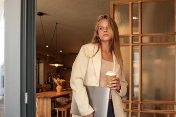 Young woman in beige blazer holding laptop and takeaway coffee cup, standing at modern cafй entrance, stylish business lifestyle concept