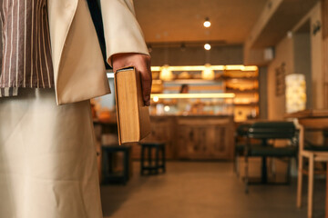 Obraz premium Close-up of woman holding old book in hand, elegant outfit with striped shirt and cream skirt, cozy modern café background with warm lights and wooden furniture