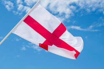 Flag of England, the St George’s Cross, waving in the wind against a bright blue sky, symbolizing...