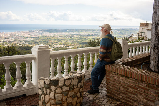 Elderly man with a white beard and green backpack sitting on a brick wall, enjoying a peaceful panoramic view of a coastal city and lush green hills under a cloudy sky. - Powered by Adobe