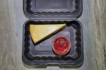 A take out container with a piece of cheesecake and disposable sauce cup. 