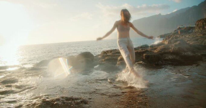 Woman dances near tide pool on rocky shore at sunset with reflection