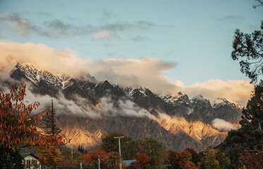 Sun hitting Queenstown snow covered mountains with clouds and a blue sky during autumn time with red and orange leaves
