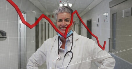 Standing doctor wearing coat and blue mask with stethoscope in hospital corridor, showing red graph