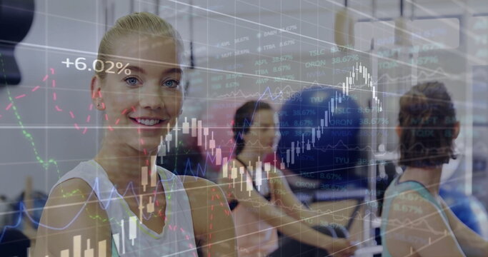 Smiling trainee wearing white tank rowing on machine in gym studio, with ball, stock chart overlay