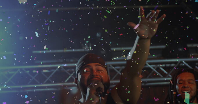 Singer wearing bandana and top holding microphone and raising hand on stage, with confetti