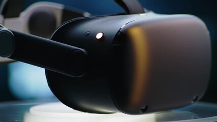A close-up, detailed shot of a modern black virtual reality headset resting on a white stand against a blurred background. - Powered by Adobe