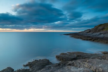 Fototapeta premium Dramatic sky over calm ocean waters reflecting coastal rocky shoreline at dusk