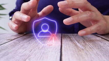 Risk in user data protection. A businesswoman's hands at a wooden desk protecting a virtual shield icon with a person avatar and a warning sign.