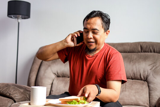Asian Man Sitting on Sofa and Talking on the Phone While Holding Plate With Sandwich - Powered by Adobe