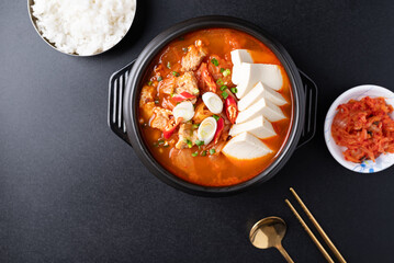 Top view of Korean food (Kimchi Jjigae), Kimchi soup with tofu, pork, scallion and chili in Korean stone bowl eating with cooked rice and kimchi on black background