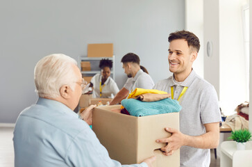 Obraz premium Helpful friendly smiling male volunteer giving senior elderly man box of second hand clothes. Volunteers at humanitarian aid center providing humanitarian help, social support, charity and donations.