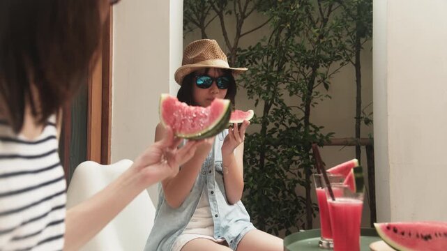 Chilling Asian family holidays, casual girl and mum in sunglasses enjoy eating fresh watermelons sliced and smoothie drinks, happiness relaxing on vacation, and hot summertime at outdoor terrace.