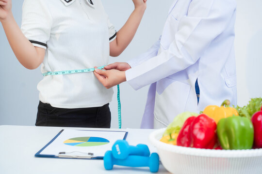 A middle-aged Asian female dietitian conducts a health consultation with a middle-aged Asian female patient at a table, focusing on weight loss, waist measurement, BMI, diet control,overall wellness
