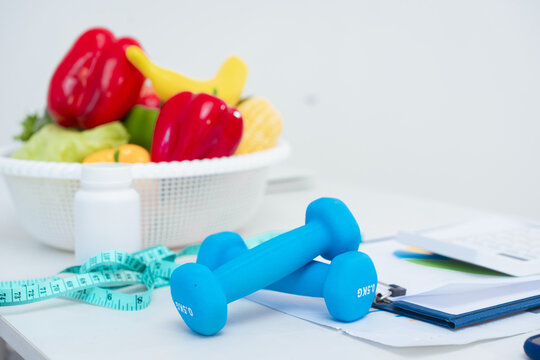 At the dining table, a nutritionist advises patients on healthy eating and weight loss. Vegetables, supplements, dumbbells, and a measuring tape are displayed, symbolizing a healthy lifestyle