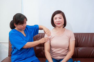 Obraz premium Young Asian nurse assisting middle-aged female patient on a couch doing physical therapy, focusing on muscle strengthening, rehabilitation, stretching,shoulder pain management in healthcare facility