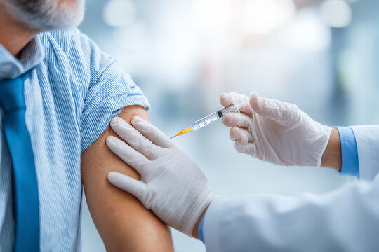 A healthcare professional wearing gloves administers a vaccine injection into the upper arm of a man dressed in a blue shirt and tie