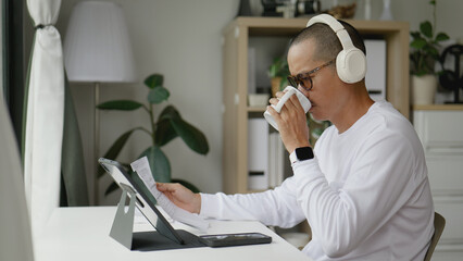 Asian man working from home on his tablet while sipping coffee.
