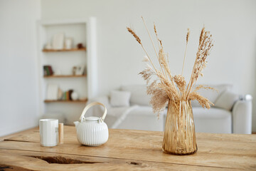 Closeup of dried wheat and grass bouquet in glass vase standing on wooden table with ceramic teapot and mug, blurred living room with sofa and bookshelf in background, copy space