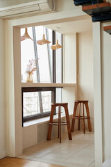 Empty home interior featuring two wooden bar stools positioned by large window with city view, dried flowers in glass vase on counter, pendant lights hanging above