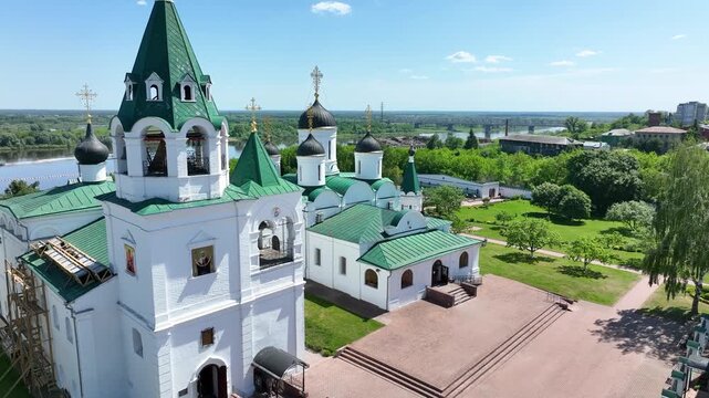 A drone view of a historic Russian Orthodox monastery with white buildings and green roofs, overlooking a river on a sunny day