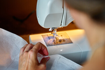 Close up of woman using modern sewing machine to stitch fabric at home under lamp light. Concept of sewing and tailoring