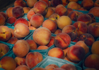 Ripe peaches are piled in blue cartons, showcasing their warm, sun-kissed hue. The fruit's fuzzy texture adds a tactile quality to the composition, evoking the feel of a bustling farmers' market. 