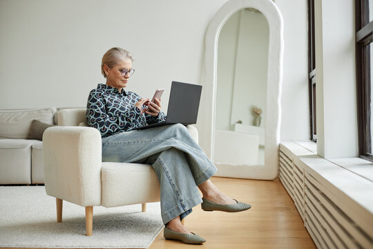 Senior Caucasian woman sitting in armchair using smartphone and laptop multitasking in modern living room, wearing glasses and looking at device screen, natural light coming from window - Powered by Adobe