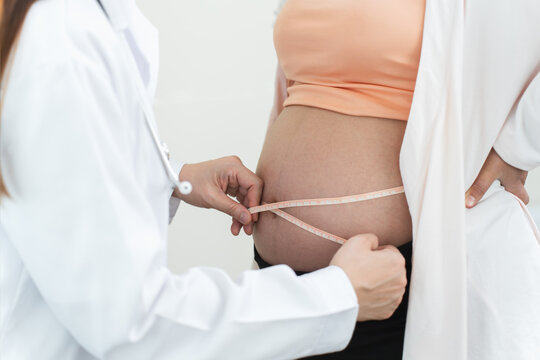 Close up of doctor hand using measuring tape to measure pregnant woman's belly in clinic. Mother in 25 weeks of pregnancy visit gynecologist for regular examining pregnancy health. Unrecognized people