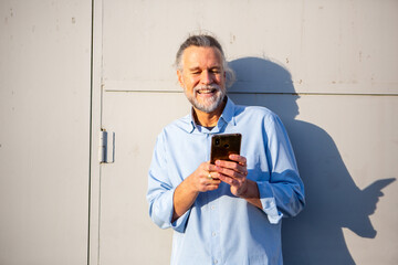 Smiling older man using smartphone while leaning on wall