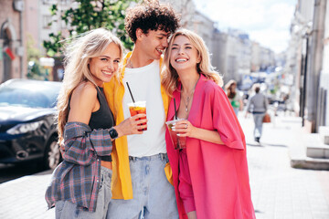 Young three stylish friends posing in the street. Fashion man and two cute female dressed in casual summer clothes. Smiling models having fun. Cheerful women and guy outdoors, Hold and drink lemonade