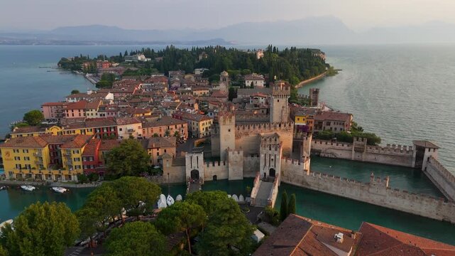 Aerial drone wide shot of Castello Scaligero di Sirmione castle and Sirmione town on Lake Garda, Italy, at summer sunrise.
