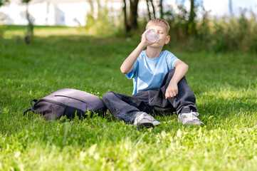 Teen boy drinking water from plastic bottle while sitting on green lawn in summer, hydration and healthy lifestyle concept