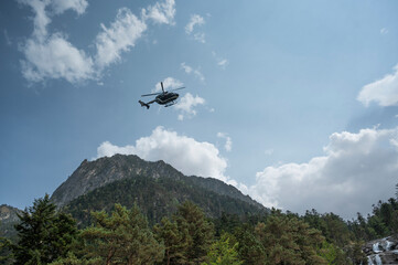A police helicopter is performing aerial maneuvers over the mountainous terrain of Cauteret in the Pyrenees, with Gavarnie visible in the background. The sky is cloudy as the helicopter hovers.