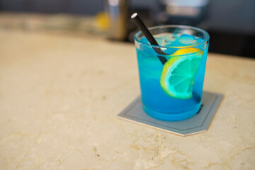 Refreshing Blue Cocktail Drink with Lemon Slice and Ice Cubes Garnished with a Black Straw Served on a Coaster at a Bar Counter