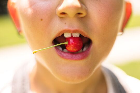 Young girl holds a red cherry between her teeth on sunny day