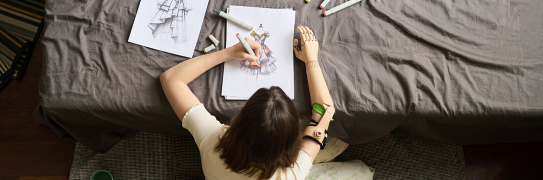 Girl with prosthesis sketching at desk in bedroom from above