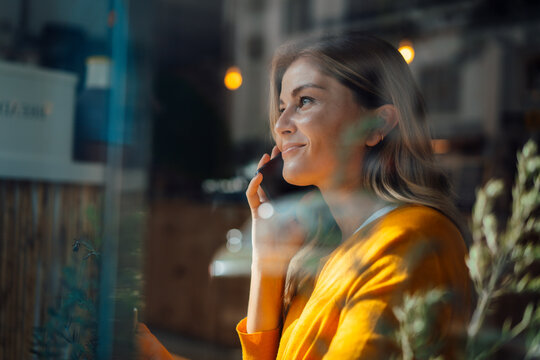 Smiling woman talking on mobile phone looking through window in cafe