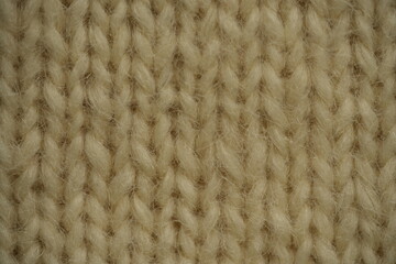Beige wool knitted fabric texture, macro photo. Close-up of soft hand-knitted wool yarn with a symmetrical pattern. Cozy, fuzzy textile surface showing detailed stitching and loop structure.
