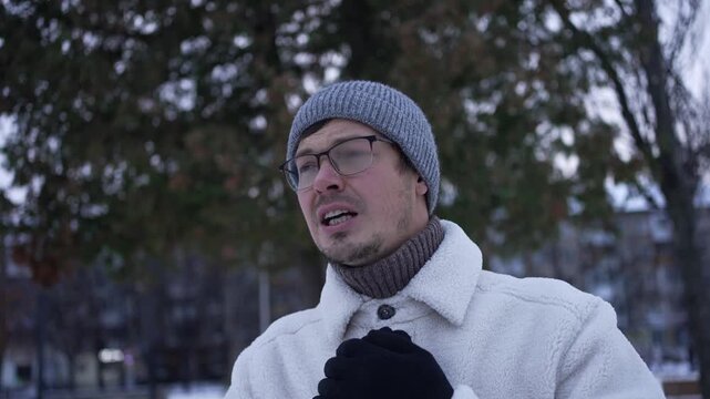 Young caucasian man wearing winter clothes, hat and gloves, is freezing and suffering from cold weather in a snowy park during winter season