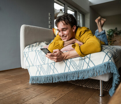 Smiling young man using smart phone lying down on sofa at home