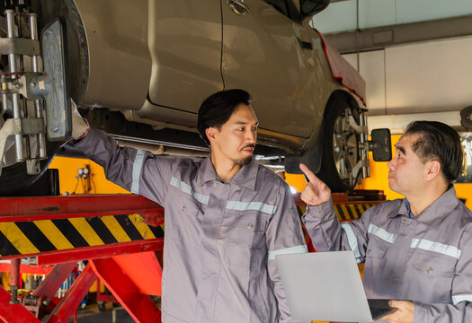 Two mechanics under car lift discussing inspection, teamwork between young and senior partner in garage small business, combining experience with modern skill in automotive maintenance service