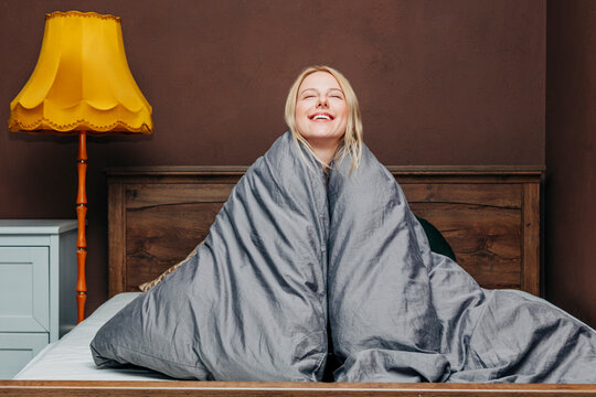 Smiling woman wrapped in gray blanket waking up in cozy bedroom