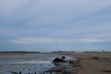 A wide, low-tide view of a sandy beach with small islands and distant structures under a cloudy sky.