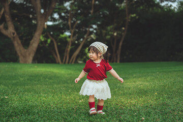 Fototapeta premium Cute baby in red outfit taking first steps on green grass in park, toddler daughter learning to walk outdoors in nature, childhood discovery and early learning concept